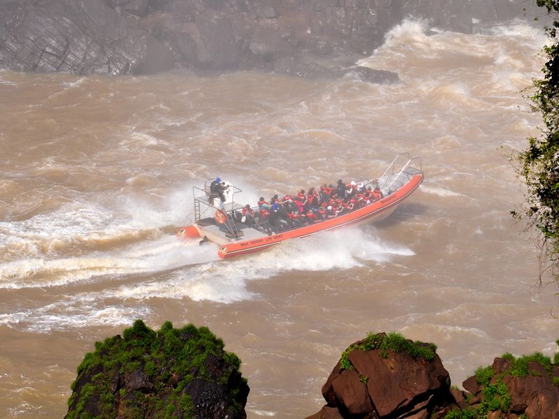 Red boat filled with passengers navigating turbulent brown water near the base of Iguazu Falls.